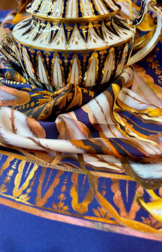 In the background of the photos a teapot that has a strong pattern of vertical feather like fronds, in Dark blue and gold on a white ground. A twill silk scarf  echoing this same pattern sits in the middle and foreground. On the scarf is a deep blue border belwo a pattern using the gold details of the fronds on the teapot. Visible in part of the gold border  are the words "Chamberlain Wordester Teapot".