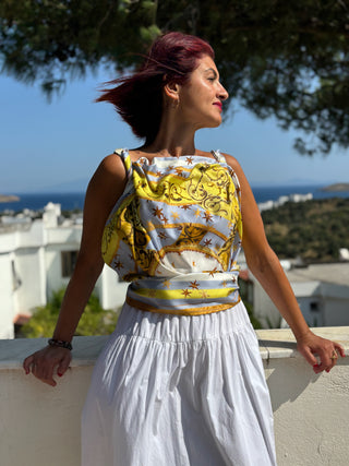 A woman wears the Lemon Starry Derby Plate Square Scarf by House of Anna Acciarini, styled as a top in golden yellow and white silk, with star motifs, standing on a sunlit terrace overlooking white houses and the sea