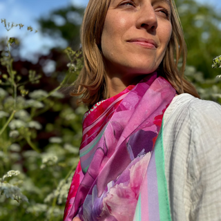 a Model wearing a designer silk scarf in pink with peony designa ndlight gerrn hand rolled edge detail in agarden inspired setting.