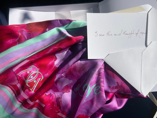 A close-up of a colorful silk scarf with a floral pattern in shades of pink, purple, and mint green, draped next to a white envelope and a handwritten card.