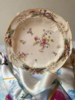 The image shows an eighteenth century porcelain plate with an arrangement of flower so in the center surrounded by, in this image four pairs of birds . The birds appear on the silk scarf behind the plate. The colour of the plate is a creamy ivory with gilding and the ground of the scarf is a lilacs powder blue.